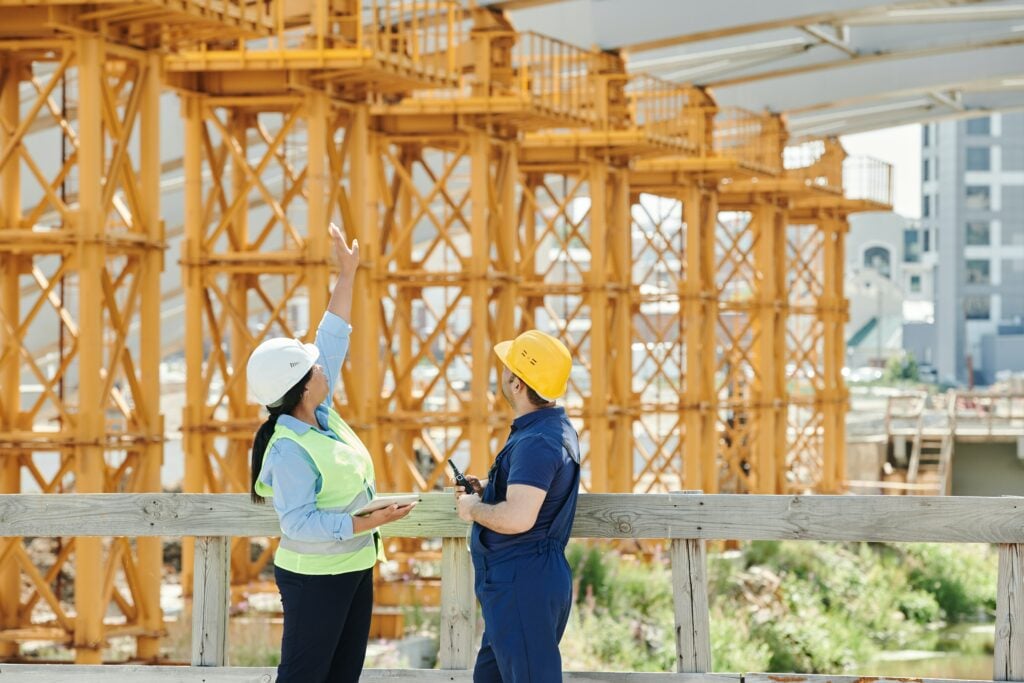 A man and woman on a construction site. The woman is pointing upwards while the man looks on.