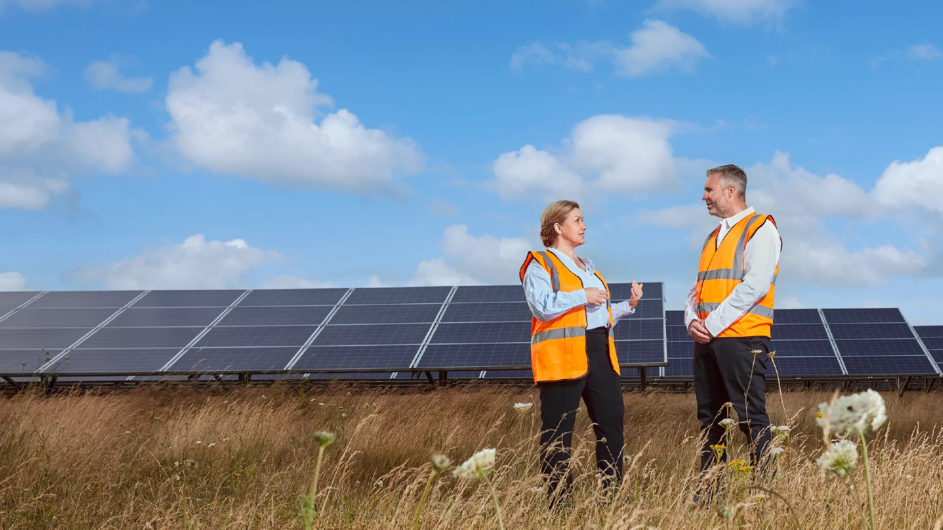 A man and a woman wearing high visibility vests speaking in front of solar panels.