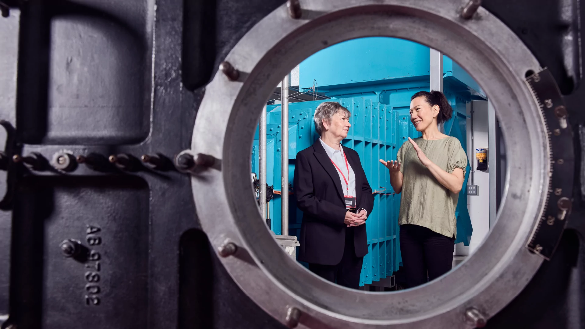 A woman and a man having a conversation seen through a porthole in a manufacturing facility.