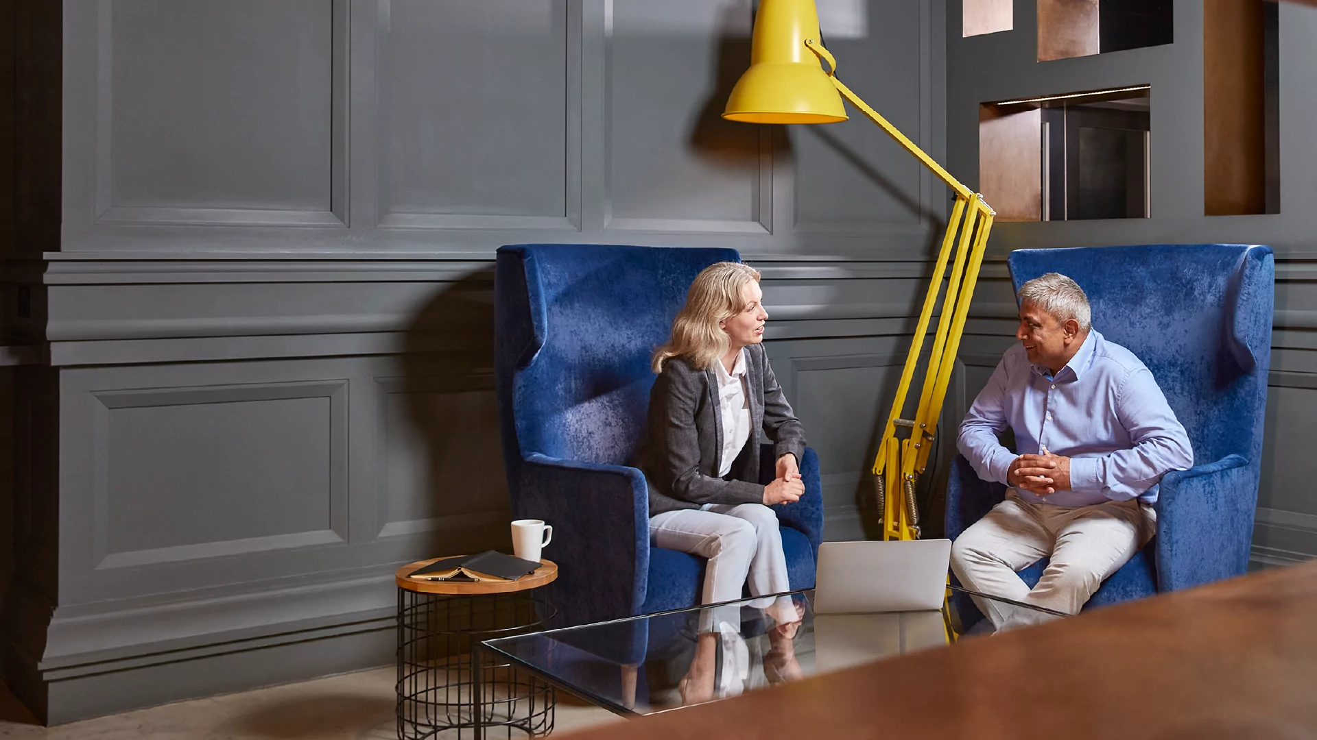A woman and a man sitting in blue armchairs under a yellow lamp, having a conversation.